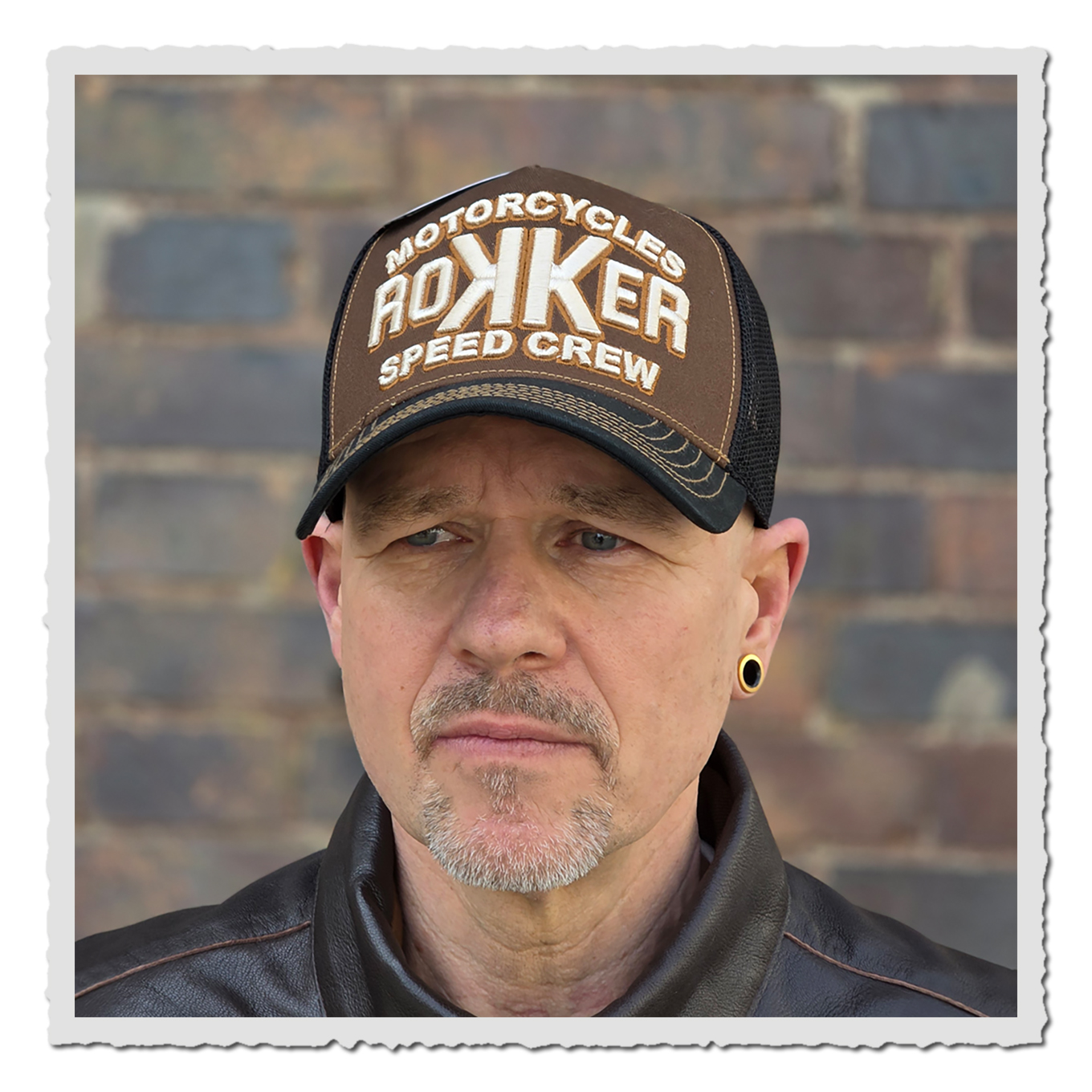 Nahaufnahme einer Person mit brauner und schwarzer Cap vor Backsteinwand, Gesicht und oberer Jackenkragen sichtbar.